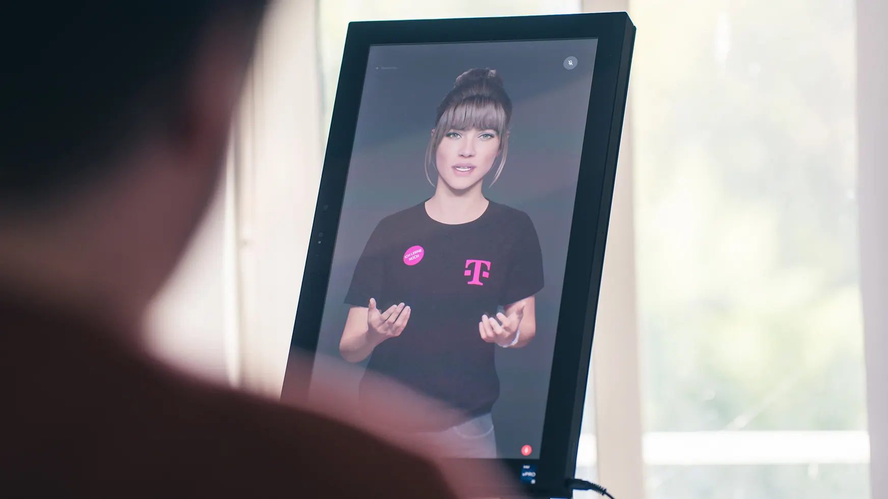 Die digitale Beraterin MIA auf einem hochkant stehenden Bildschirm im Telekom-Shop, im Vordergrund unscharf ein Kunde.