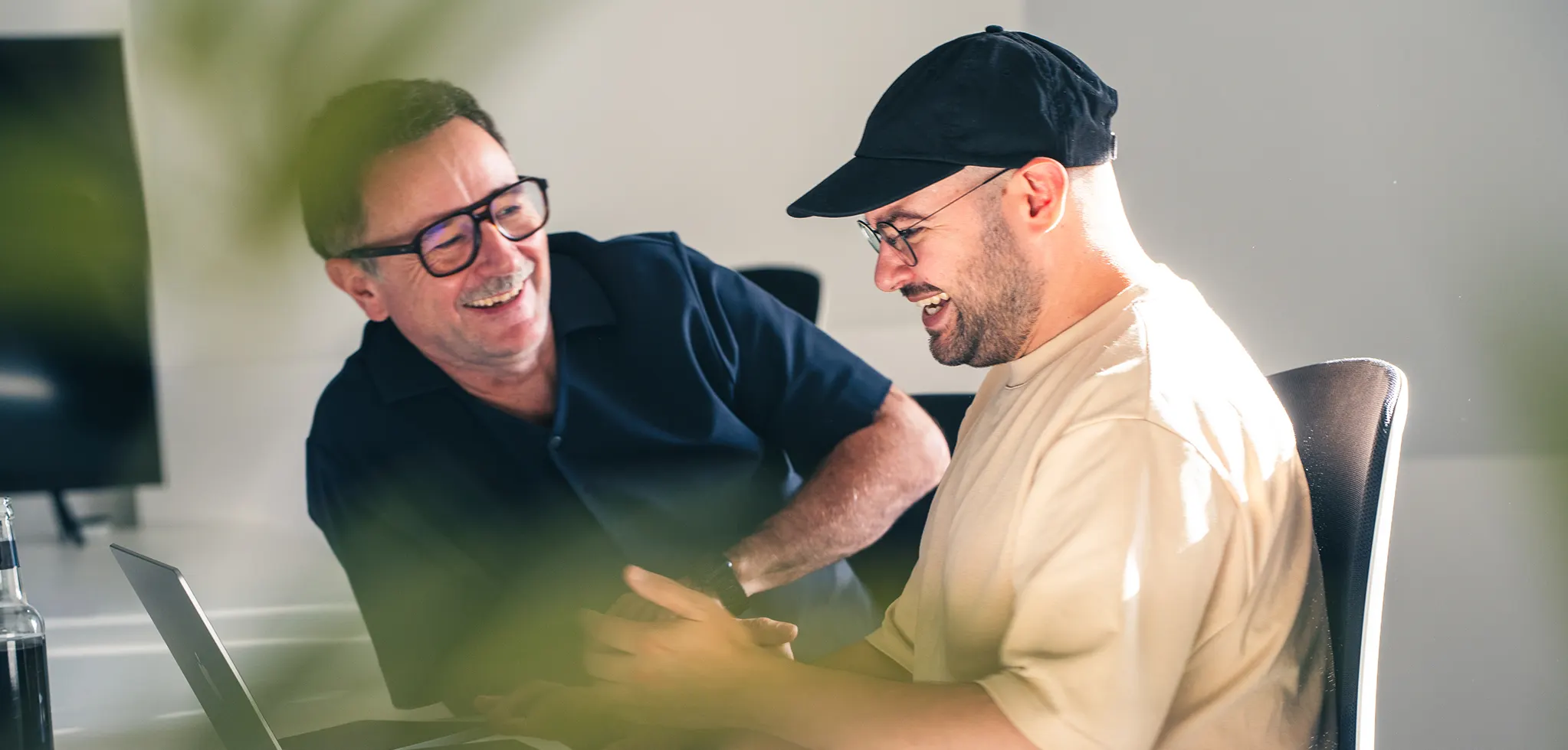 Zwei Kollegen lachen gemeinsam am Schreibtisch über einen Laptop.